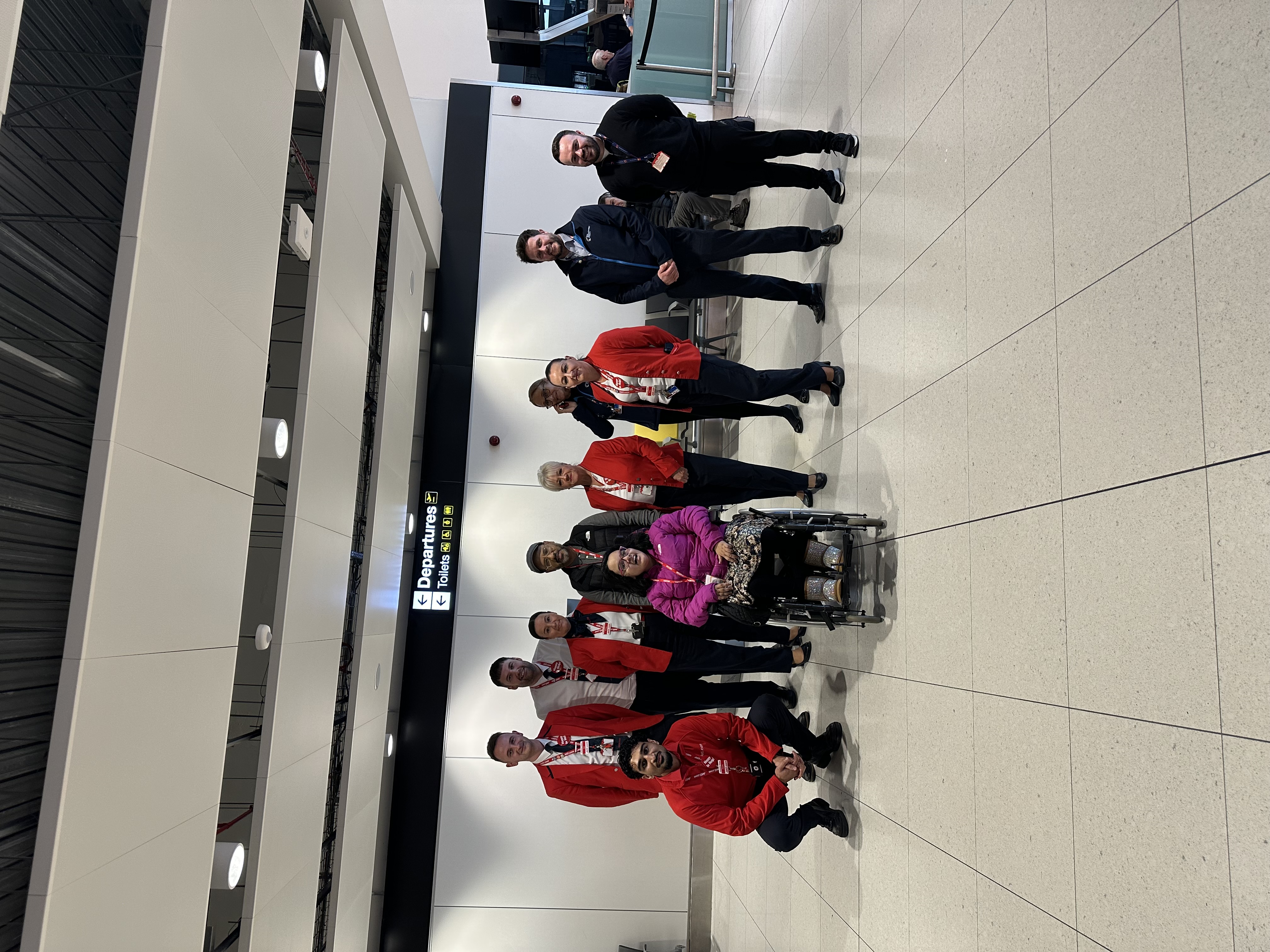 A group of airport staff in red uniforms poses with a woman in a wheelchair at Manchester Airport's departures area.