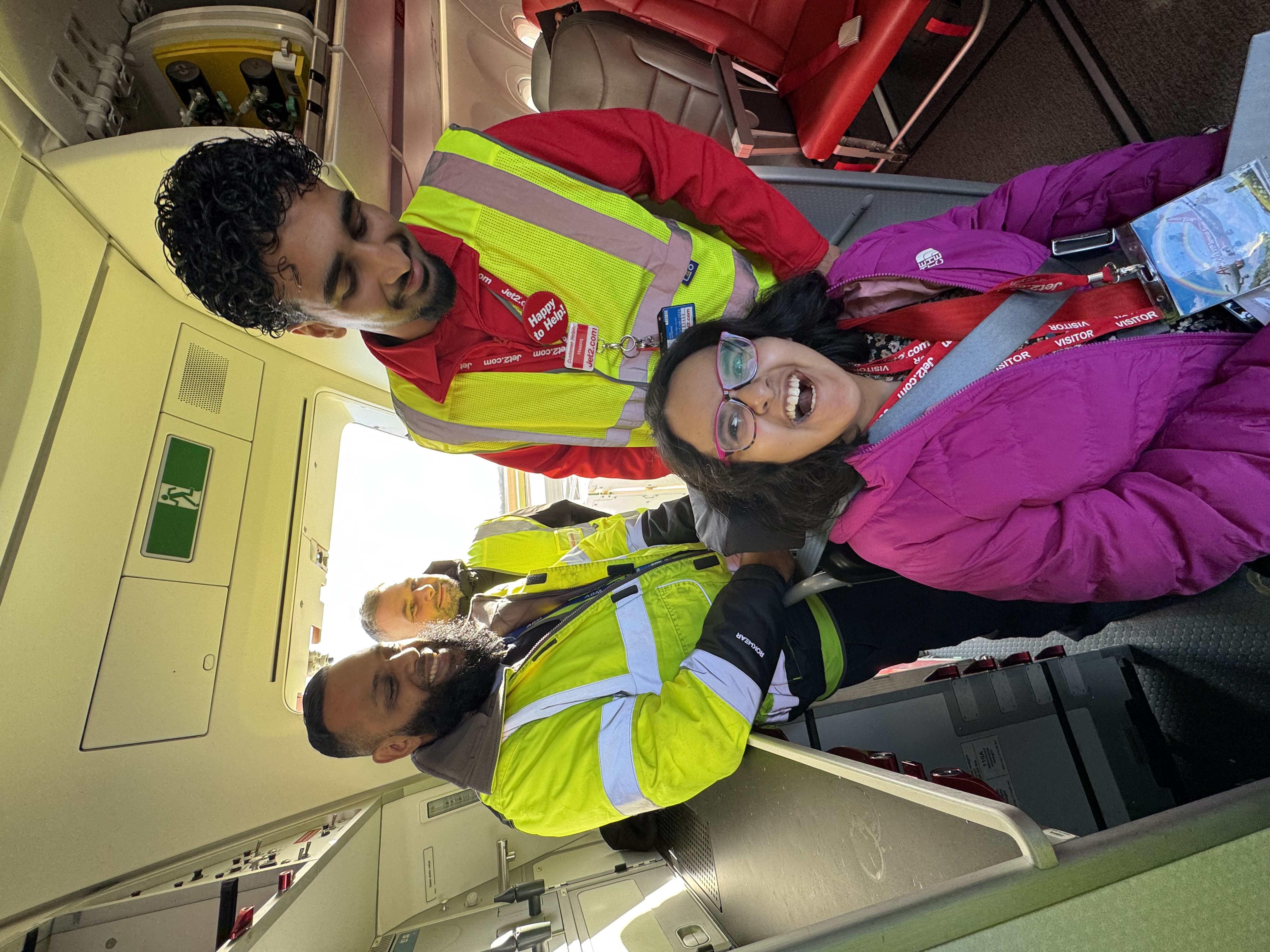 A smiling young girl in a pink coat sits in an airplane cabin, surrounded by three staff members in high-visibility jackets.