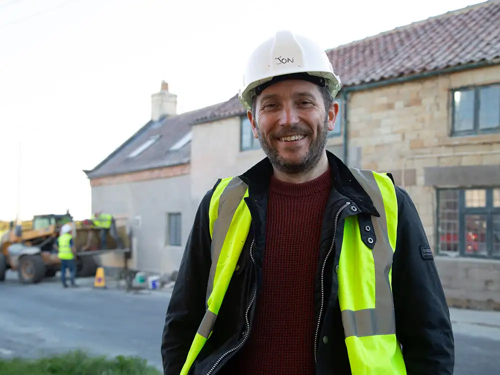 Jon Richardson in a hard hat and high-visibility jacket, smiling in front of The Plough pub in Fadmoor during renovations.