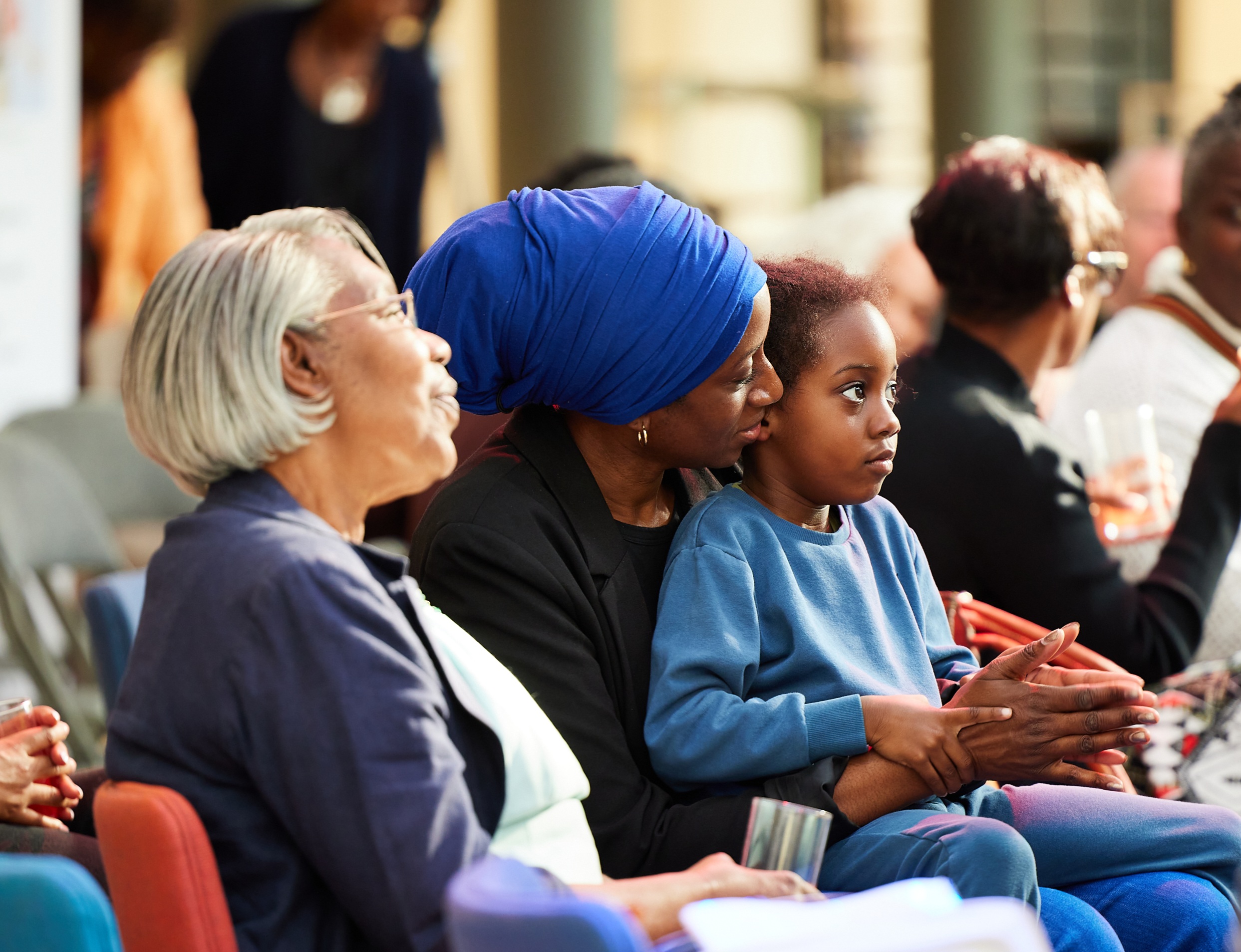 A woman in a blue headscarf holds a child, surrounded by an audience of diverse adults, in a cozy indoor setting.