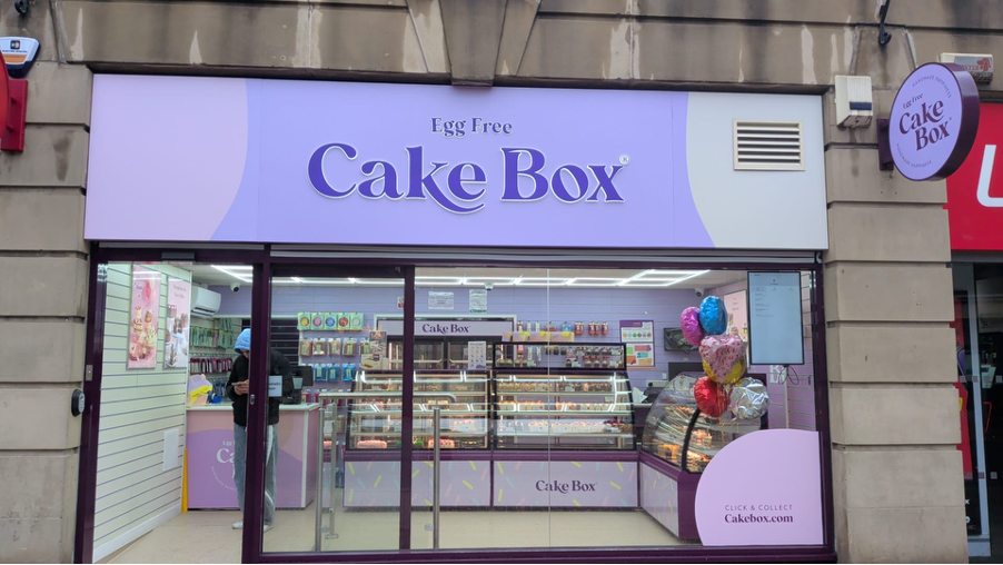 A pastel purple cake shop with colorful balloons, showcasing a variety of cakes inside, located in Yorkshire.