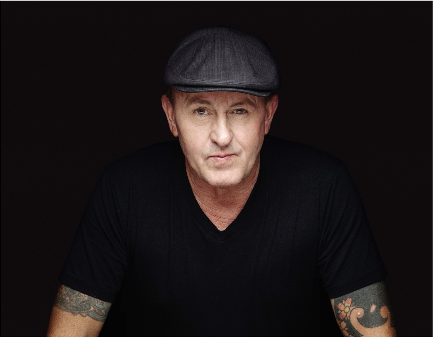 Close-up portrait of a man with a flat cap and tattoos, exuding a confident and relaxed demeanor against a dark background.