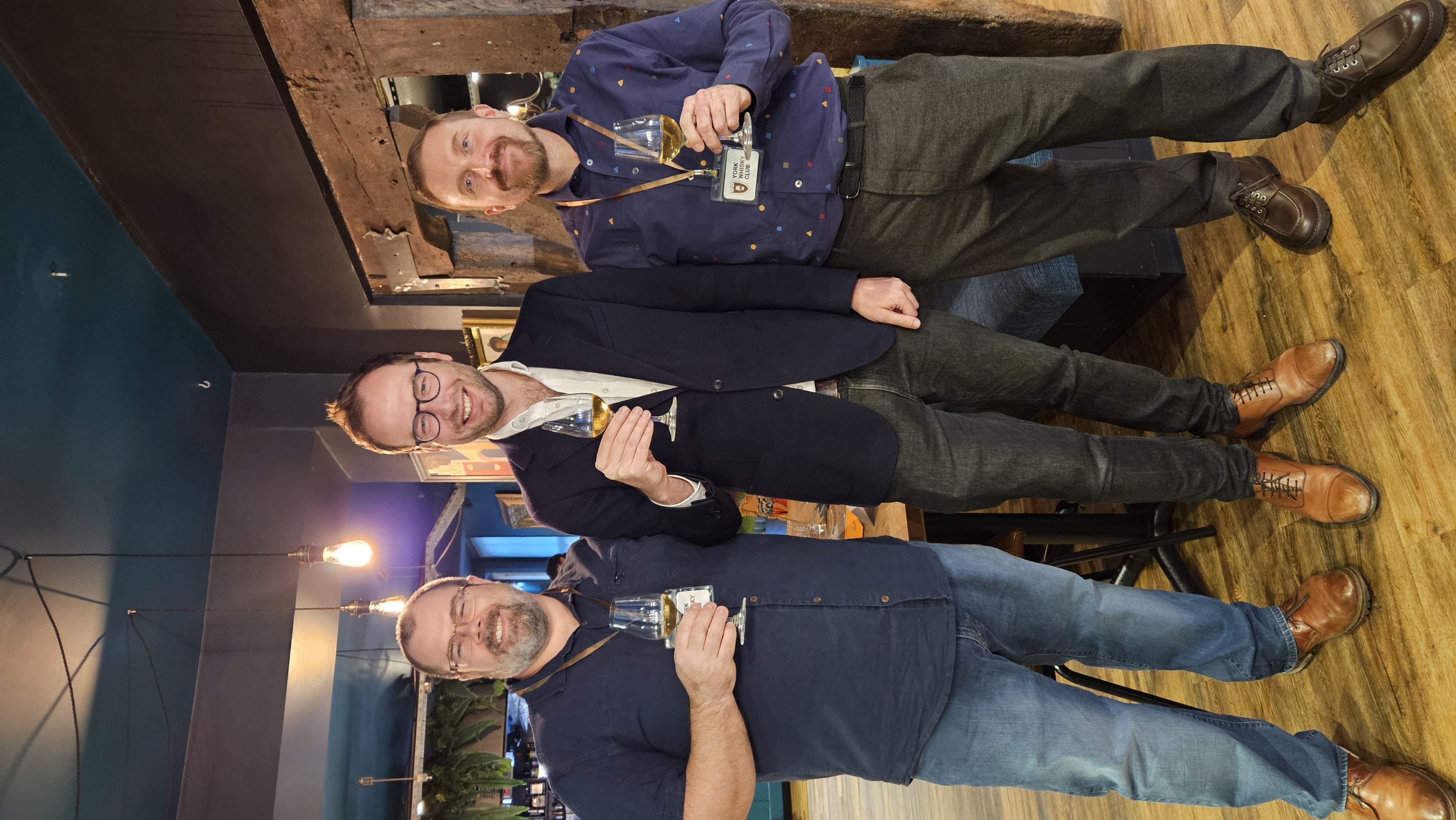 Three men in smart casual attire hold drinks, smiling in a cozy, stylish bar setting with warm lighting and rustic decor.