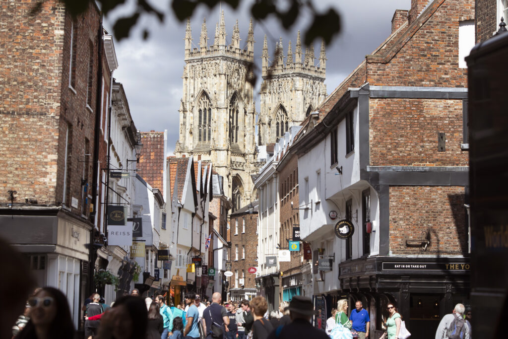 Bustling street in York with historic buildings, shoppers, and the towering York Minster in the background under a cloudy sky.