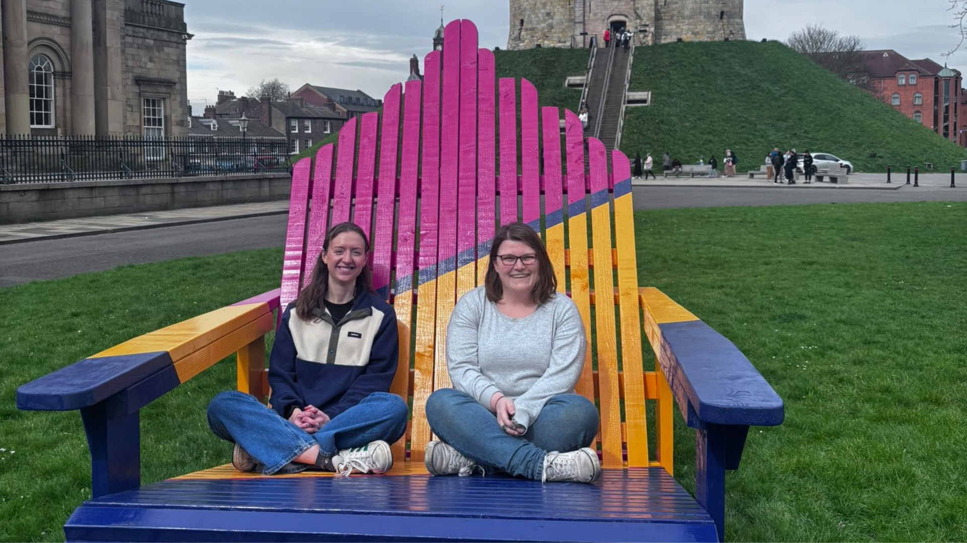 Two women sit on a large, colorful deck chair in York, with a grassy hill and historic buildings in the background.