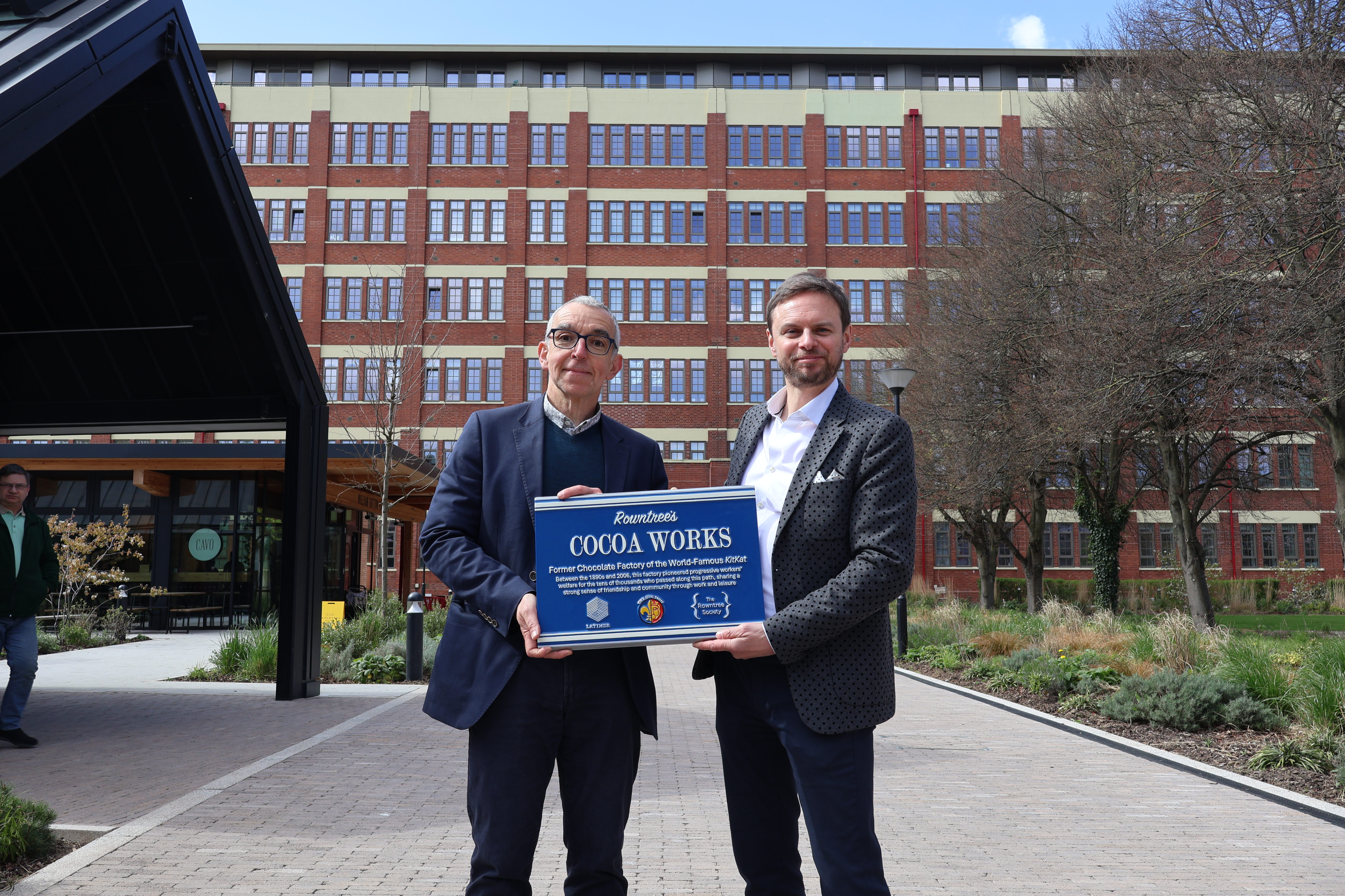 Two men hold a blue plaque reading "Cocoa Works" in front of a large red-brick factory building in York.