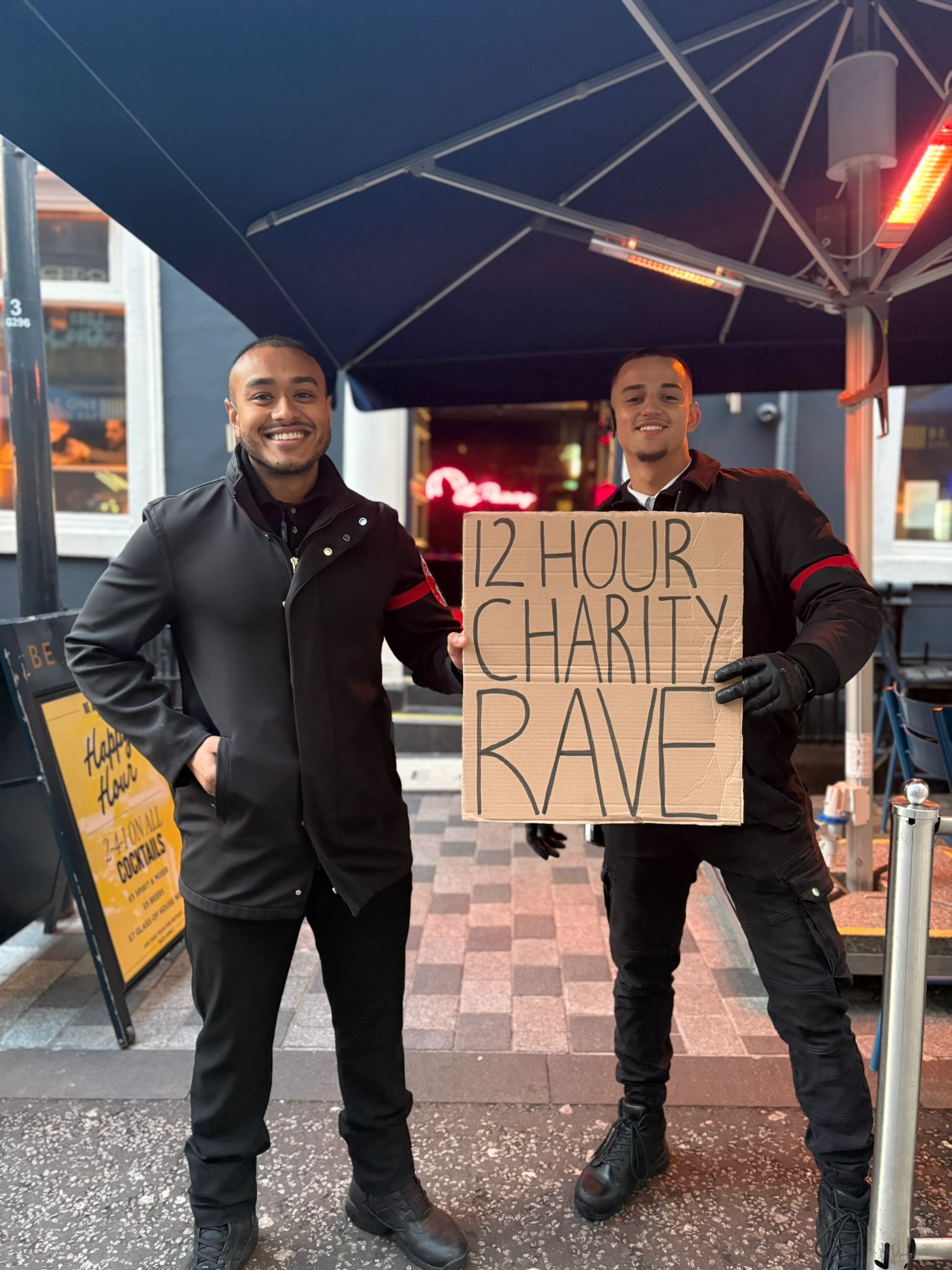 Two people holding a 12-hour charity rave sign under a blue umbrella