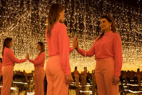A woman in a pink outfit touches her reflection in a mirrored room adorned with warm glowing lights.