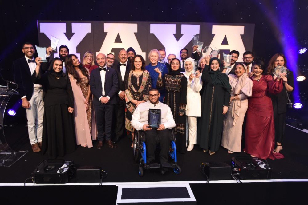 A diverse group of award winners poses proudly on stage, surrounded by balloons and bright lights, celebrating their achievements.