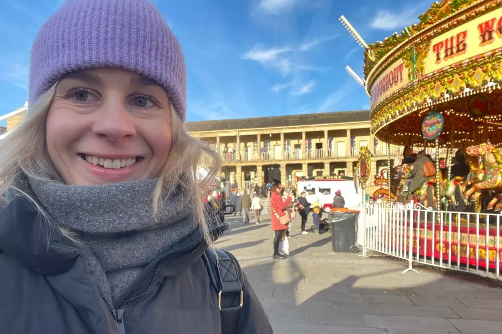 A smiling woman in winter attire stands near a vibrant carousel at a festive market with blue skies above.