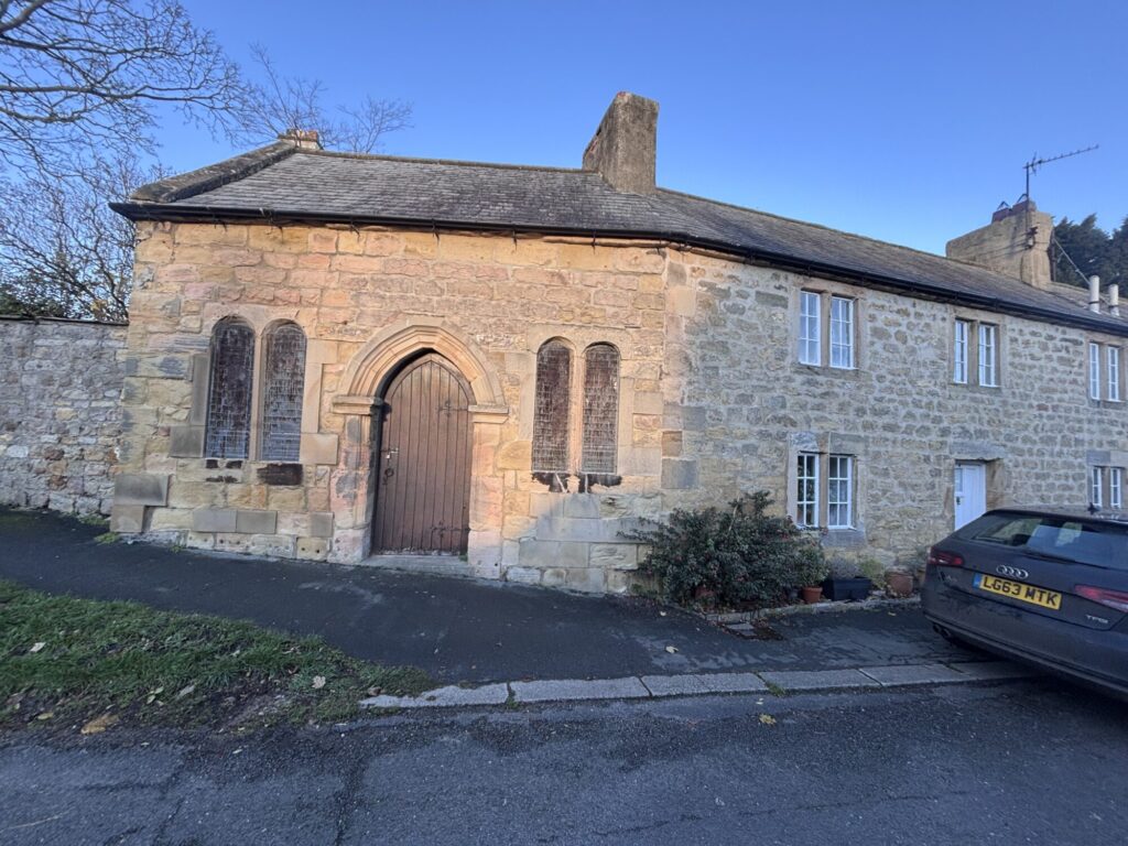 Historic stone building with an arched wooden door, large windows, and a low stone wall, set on a quiet street.