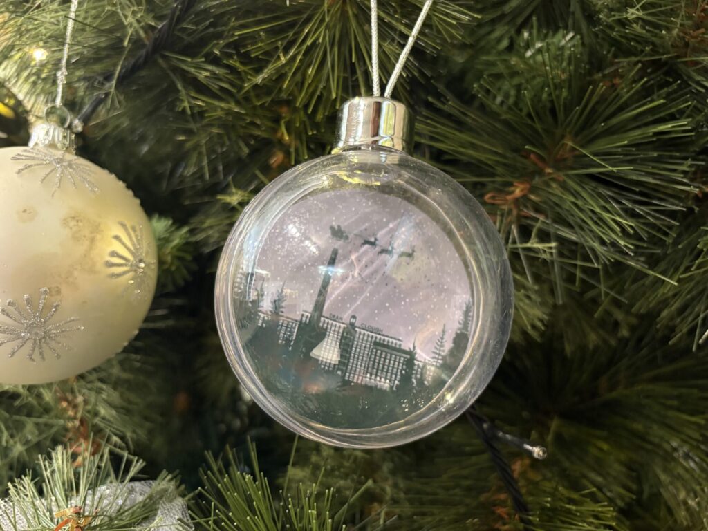 A clear Christmas ornament hangs from a pine tree, featuring a silhouette of a scene with snow and flying birds inside.