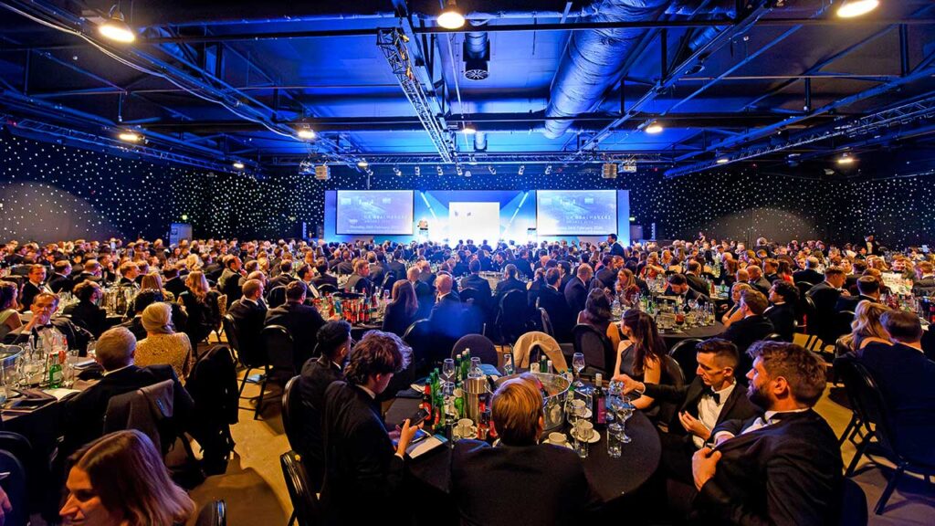 A packed event venue filled with attendees in formal attire, illuminated by blue lighting and decorated with starry backdrops.
