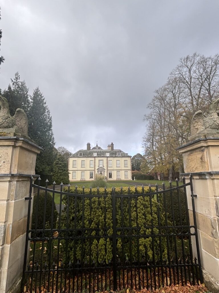 Grand Myton Hall seen through ornate black gates, surrounded by manicured greenery and under a cloudy sky.