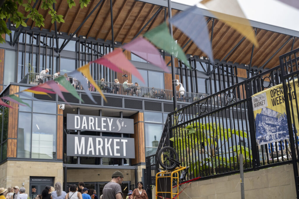 Colorful bunting hangs in front of Darley Street Market, bustling with visitors on a sunny day. Modern architecture visible.