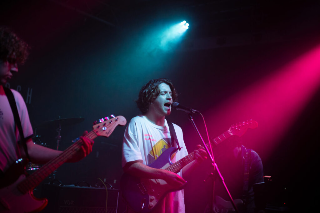 A musician with curly hair sings passionately into a microphone while playing an electric guitar, bathed in vibrant blue and pink lights.