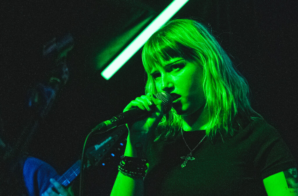 A singer with green-tinted lighting passionately performs into a microphone, showcasing vibrant energy on stage.