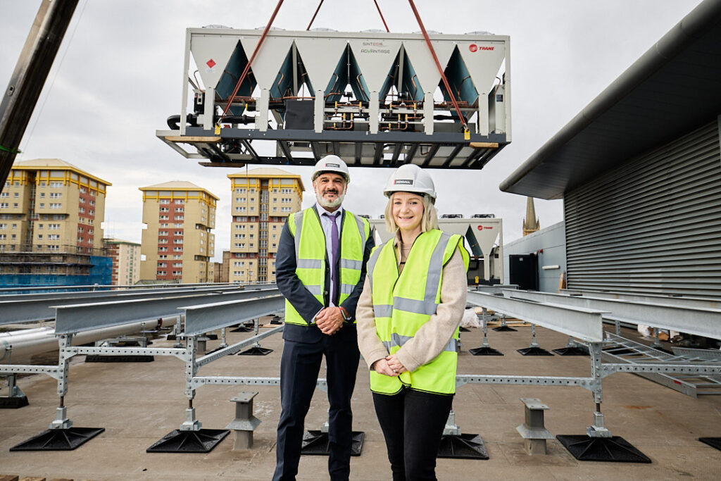 Two individuals in safety vests and helmets stand on a rooftop with a large air conditioning unit being installed in the background.