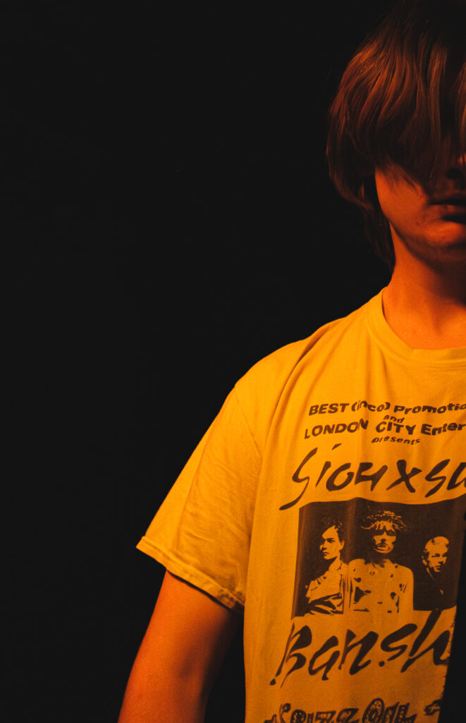 A person in a yellow graphic t-shirt stands against a dark background, partially obscured by their hair.