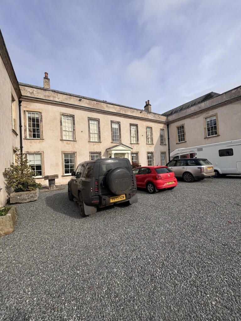 Historic building with a light-colored façade, surrounded by cars in a gravel courtyard under a cloudy sky.