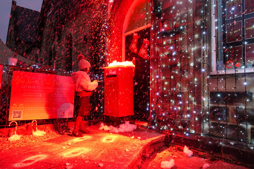 A child in a winter hat stands by a bright red mailbox, surrounded by festive lights and artificial snow.