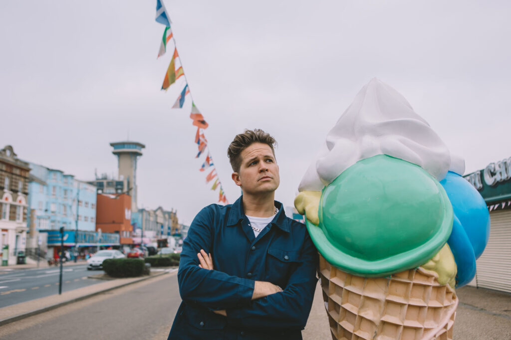 A man with crossed arms stands beside a large ice cream cone sculpture, colorful flags and a tower in the background.