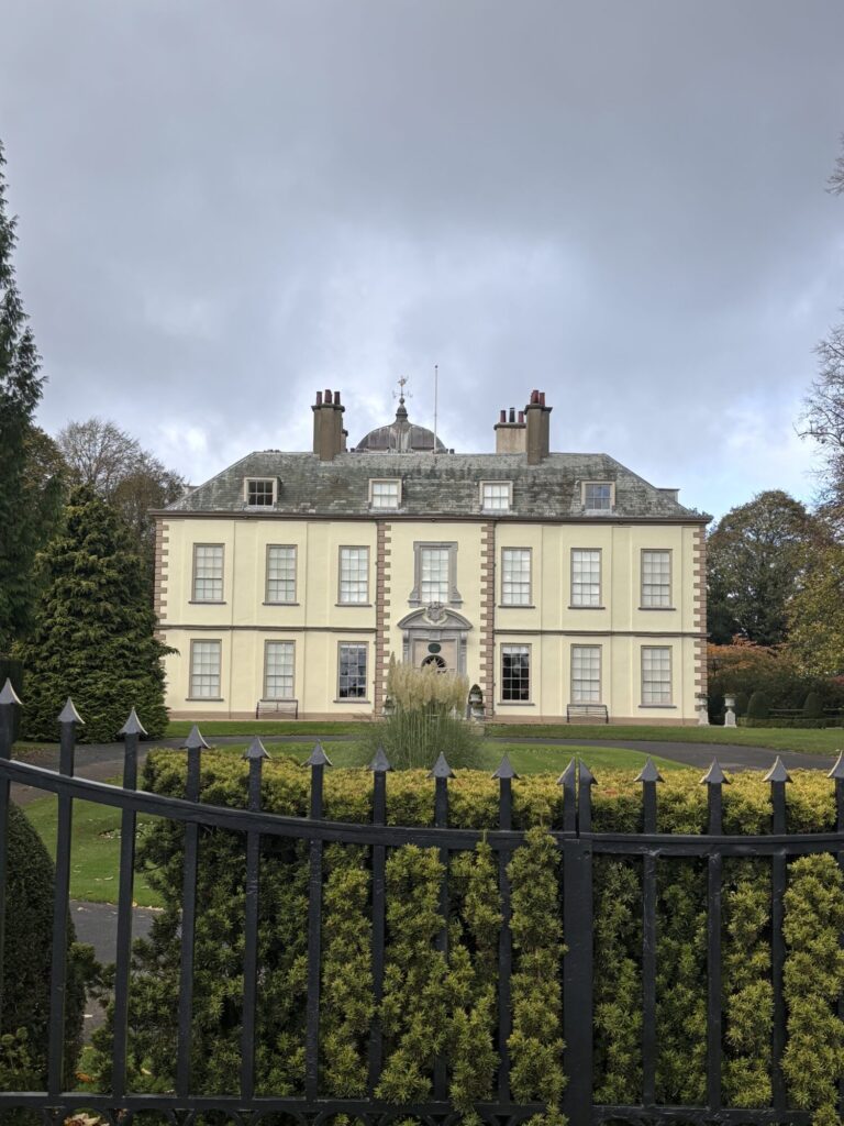 A grand historical building with a symmetrical facade, surrounded by manicured gardens and tall hedges under a cloudy sky.