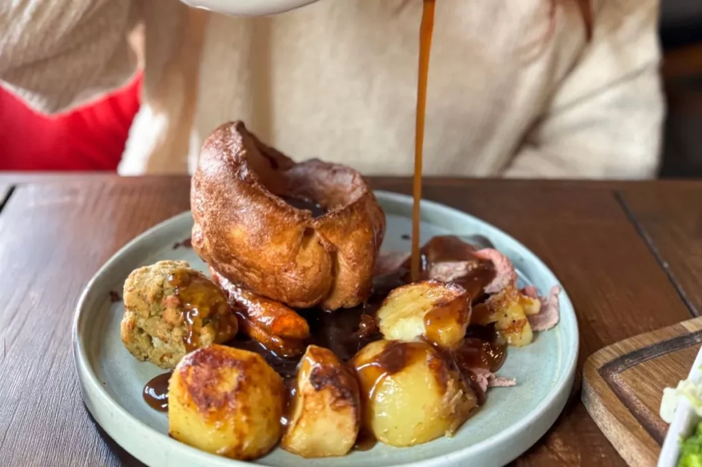 Golden Yorkshire pudding sits on a plate surrounded by roast potatoes and gravy, with a hand pouring sauce on top.