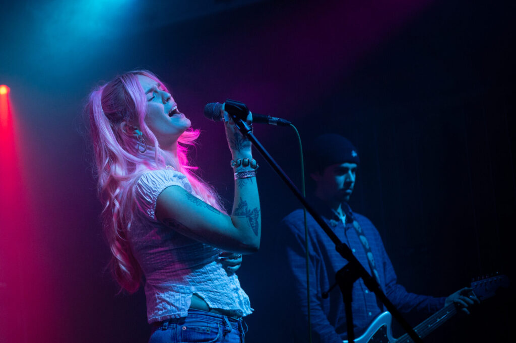 A singer with pink hair passionately performs on stage, lit by vibrant blue and pink lights, while a guitarist stands nearby.