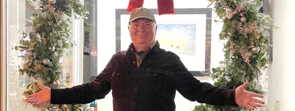 A man in a black jacket and hat smiles, standing between decorated festive wreaths with silver ornaments.