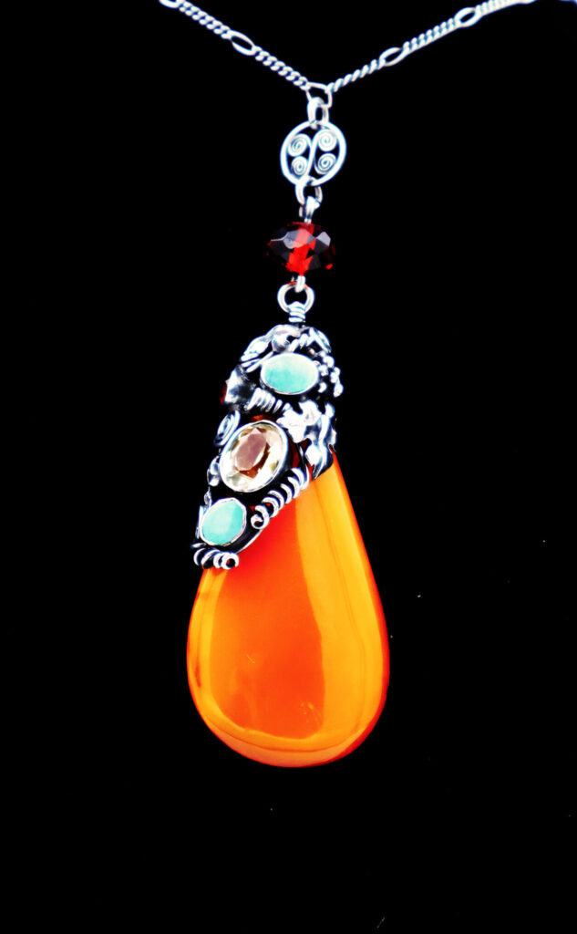 Vibrant orange teardrop pendant adorned with silver accents and colorful gemstones, set against a black background.