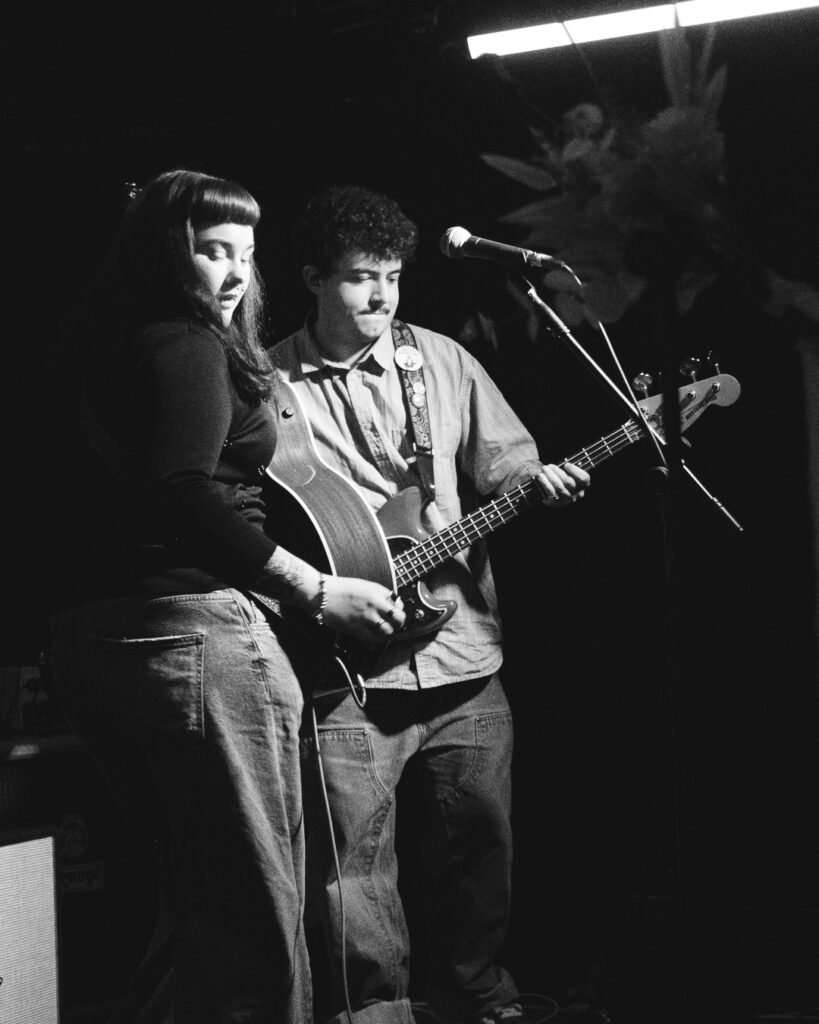 Two musicians perform on stage: one plays guitar, the other holds a bass, illuminated by stage lights in a dark venue.