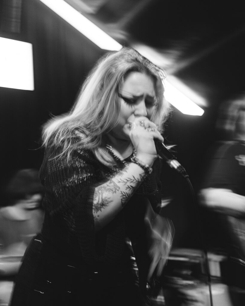 A singer with long blonde hair passionately performs on stage, blurred lights in the background enhance the energy.