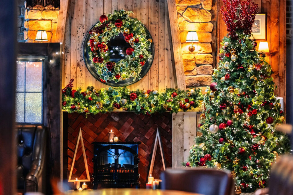 Festive interior featuring a decorated Christmas tree, a wreath on the wall, and a cozy fireplace with greenery and candles.