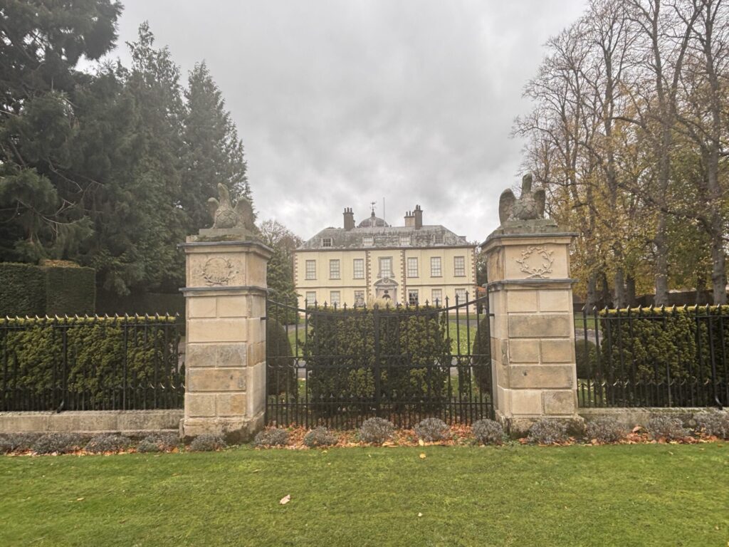 Elegant yellow Myton Hall behind ornate stone gates, framed by lush greenery and tall trees under a cloudy sky.