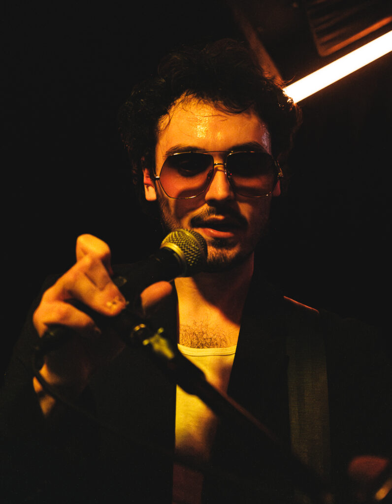 A musician with sunglasses holds a microphone, illuminated by warm, dim lighting, exuding a passionate performance vibe.
