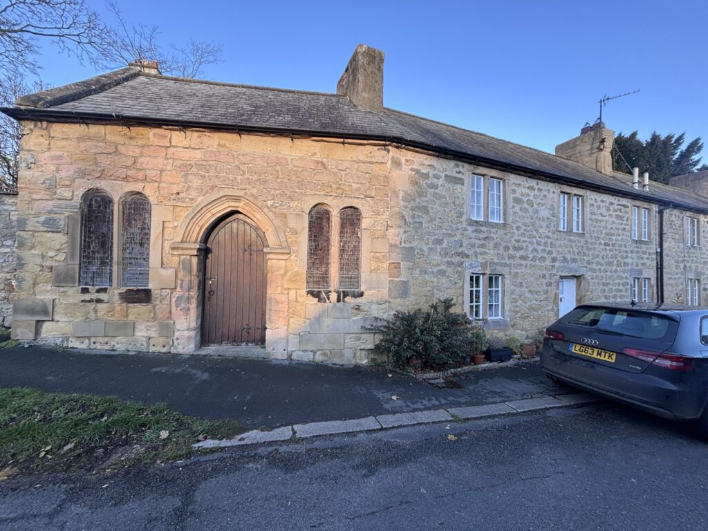Historic stone cottages with arched windows, a wooden door, and a car parked on a quiet street in Well, Yorkshire.