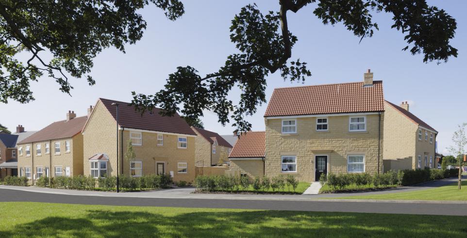 Two rows of charming yellow stone houses with red roofs lined by green lawns and leafy trees under a clear blue sky.
