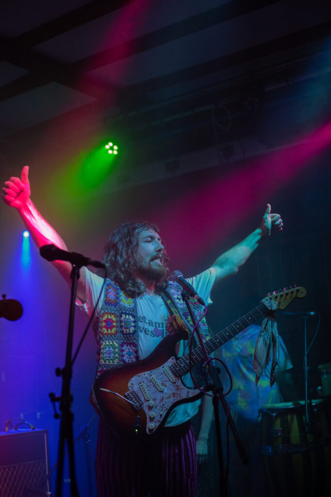 A musician in a brightly patterned vest performs on stage, raising arms in excitement amid colorful stage lights.