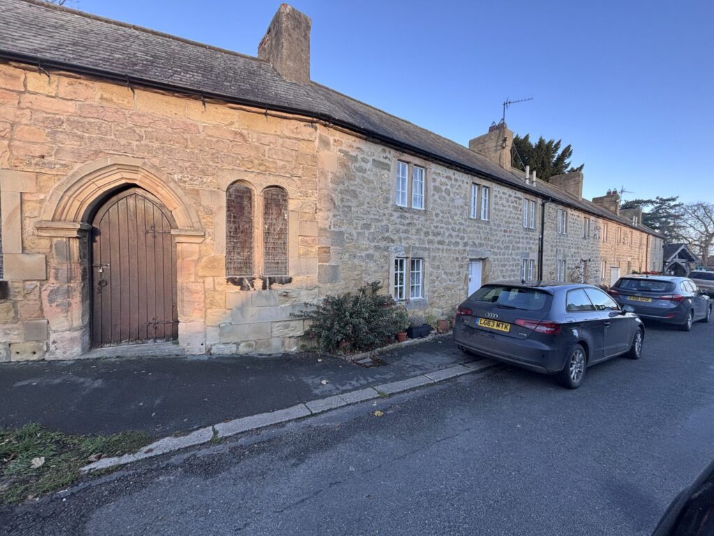 Historic stone cottages line the street, with a charming arched doorway and vintage vehicles parked nearby under a clear sky.