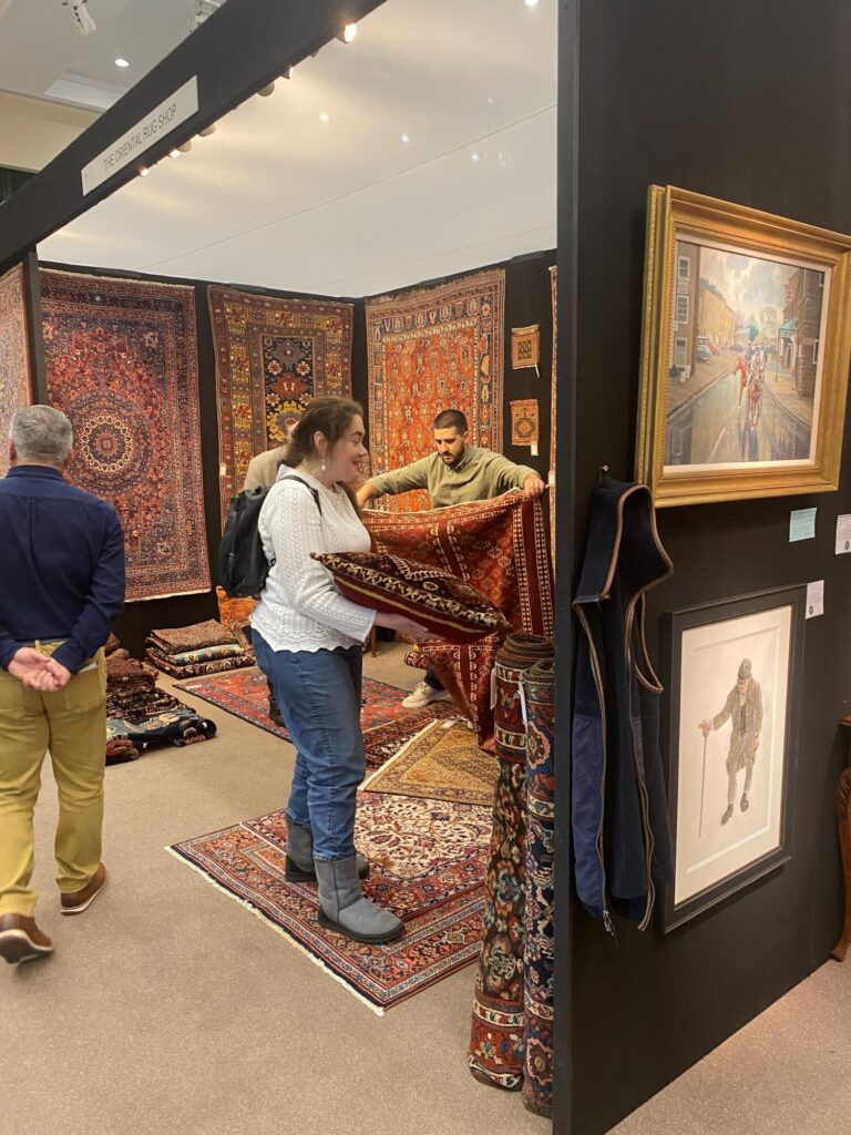 A woman inspects a vibrant rug while a man adjusts another in a well-curated antiques booth at the fair.