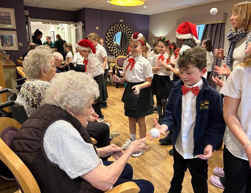 Schoolchildren in festive attire perform for elderly residents, sharing smiles and joy in a cozy care home setting.