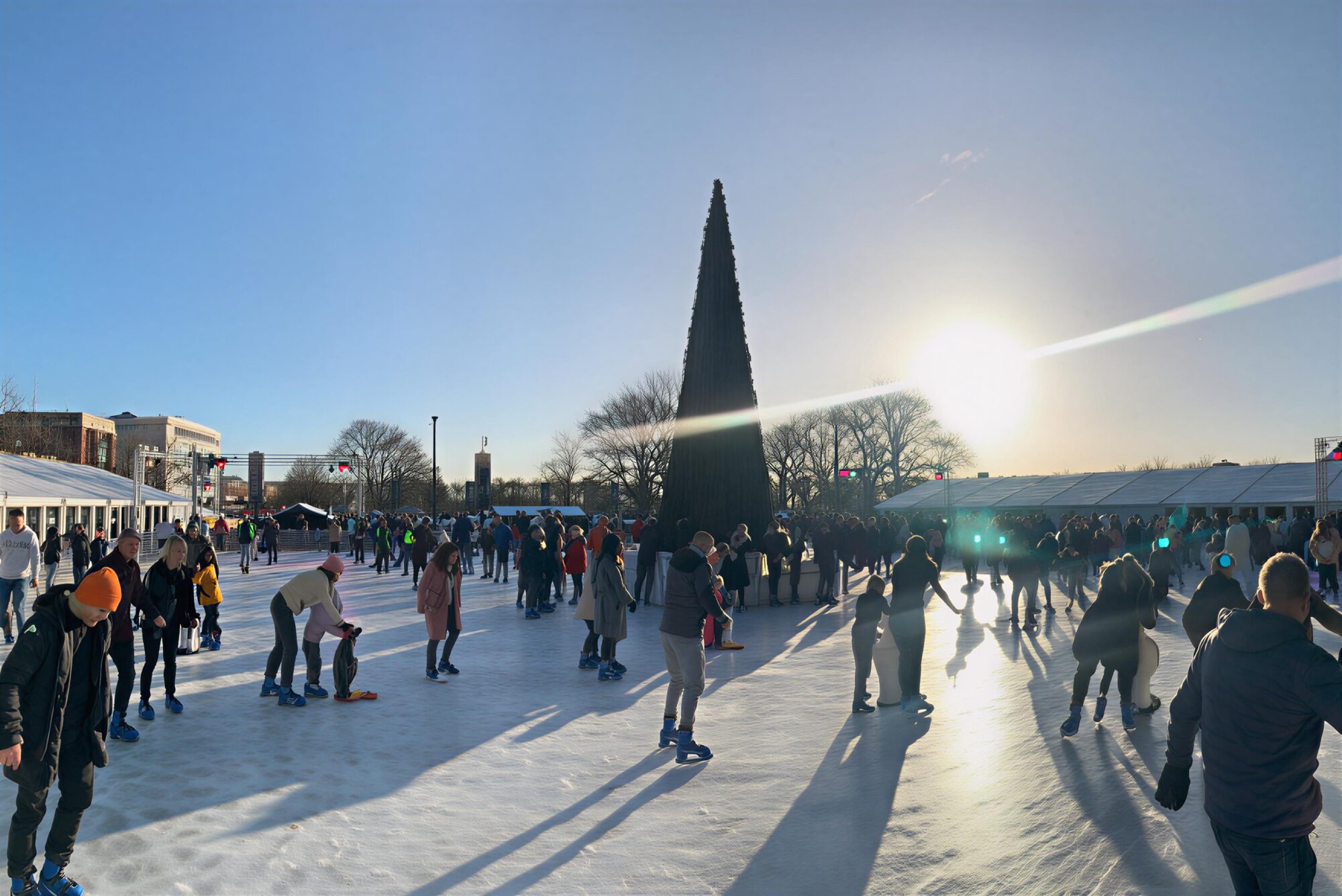 Skaters glide on ice amid a festive atmosphere, with a tall Christmas tree and bright sun in the background.