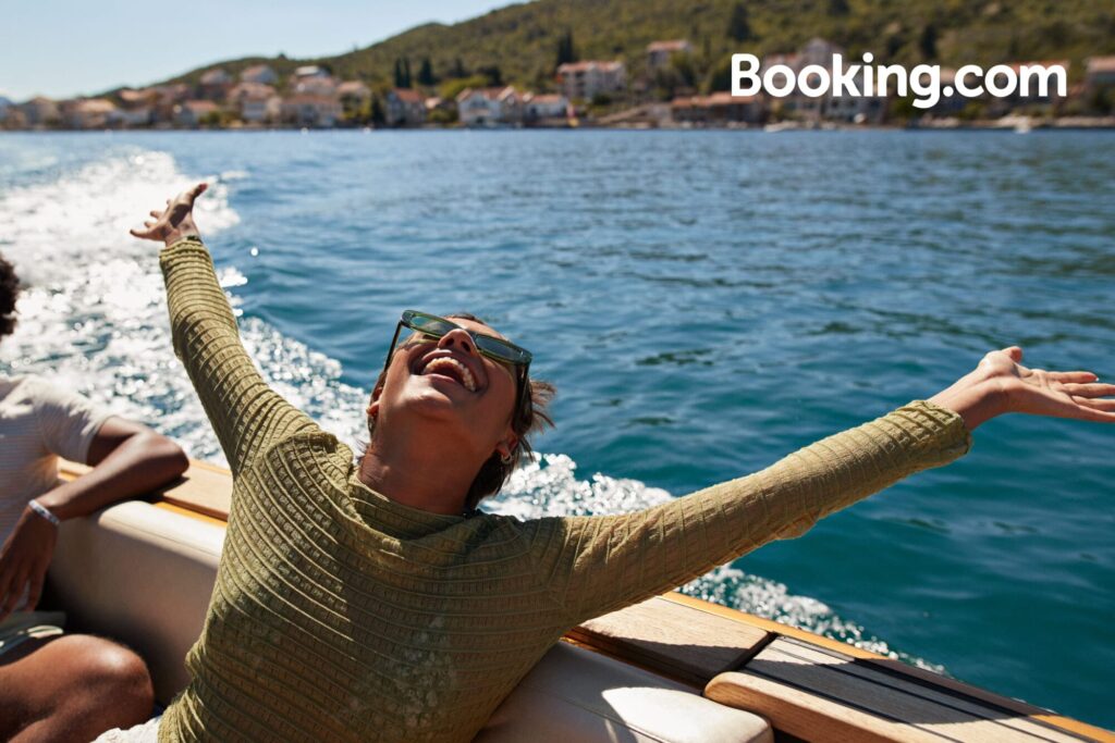 Woman with outstretched arms smiles joyfully on a boat, water glistening in the background with hills and houses.