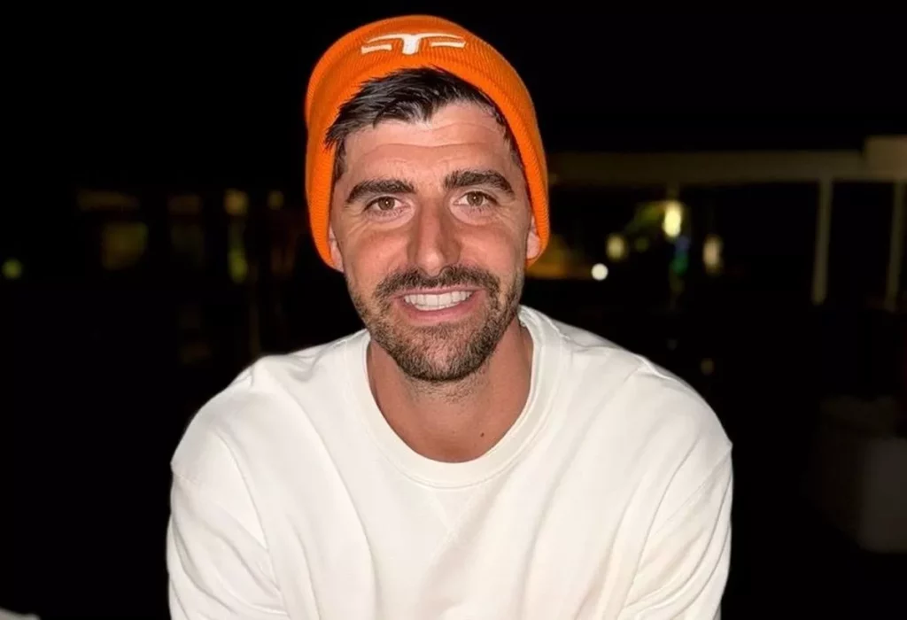 Smiling man in an orange beanie and white sweatshirt, sitting in a dimly lit setting.