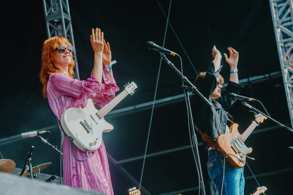 Two musicians on stage, one with an electric guitar and the other clapping, both wearing stylish outfits and sunglasses.