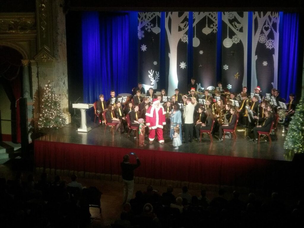 A festive stage with Santa and performers, surrounded by musicians in holiday attire, decorated trees, and blue backdrops.