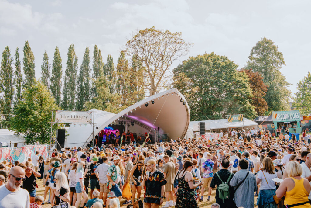 A vibrant festival scene with a large crowd enjoying live music under a sunny sky, surrounded by trees and tents.
