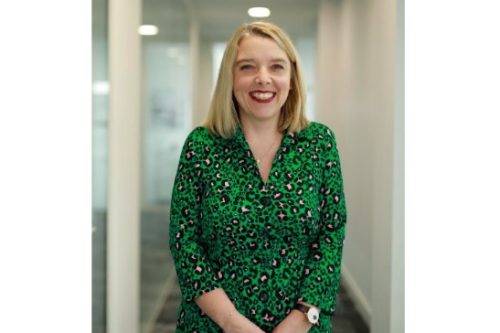 A woman with blonde hair smiles, wearing a vibrant green patterned blouse in a modern office setting.