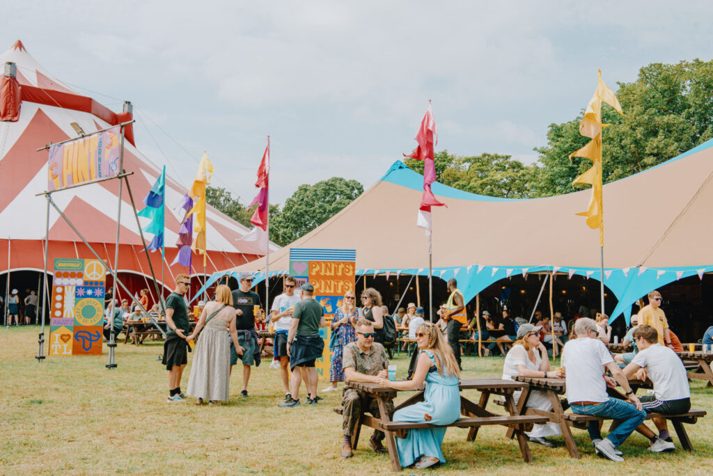 Colorful tents and flags decorate a festival scene, with people enjoying drinks and relaxing on picnic tables.